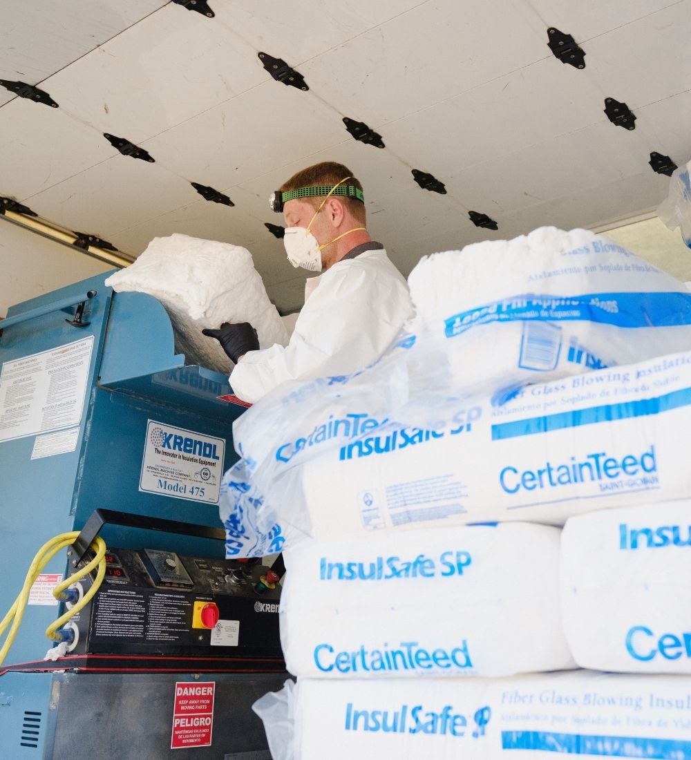 A worker wearing protective gear loads insulation material into a machine. In the foreground are several bags labeled “CertainTeed InsulSafe SP.” The worker is inside a truck or workspace with insulation supplies.