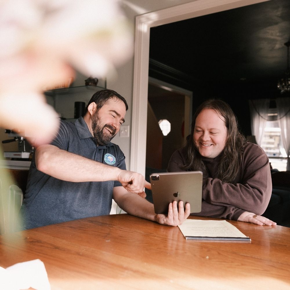 Two people sitting at a wooden table, smiling and looking at a tablet together. One person is pointing at the screen, while the other holds the tablet. A notebook lies on the table. The setting appears to be a cozy home.