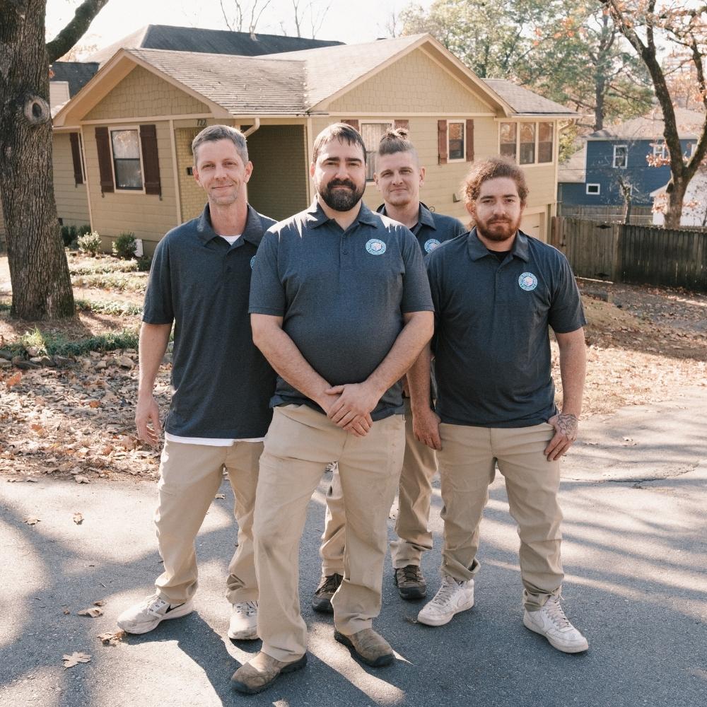 Four men wearing matching gray polo shirts and khaki pants stand together on a suburban street in front of a tan house with trees and fallen leaves in the background.