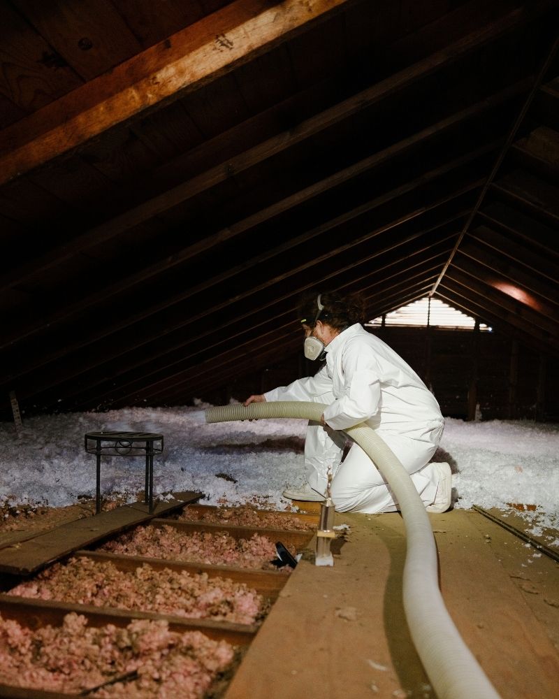 A person in protective gear and a mask kneels in an attic, using a hose to blow loose insulation onto the attic floor. Sunlight filters through a small window, illuminating the wooden beams and insulation material.