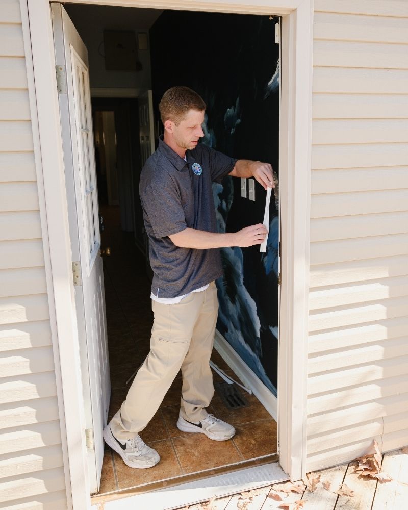 A man stands in a doorway measuring the width with a tape measure. The door is open, and he is focused on taking the measurement inside a house with tiled flooring and light-colored siding outside.