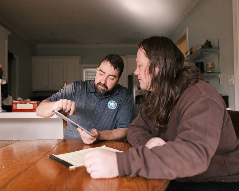 Two people sitting at a wooden table in a kitchen discuss paperwork; one man points at a tablet while the other listens and takes notes.