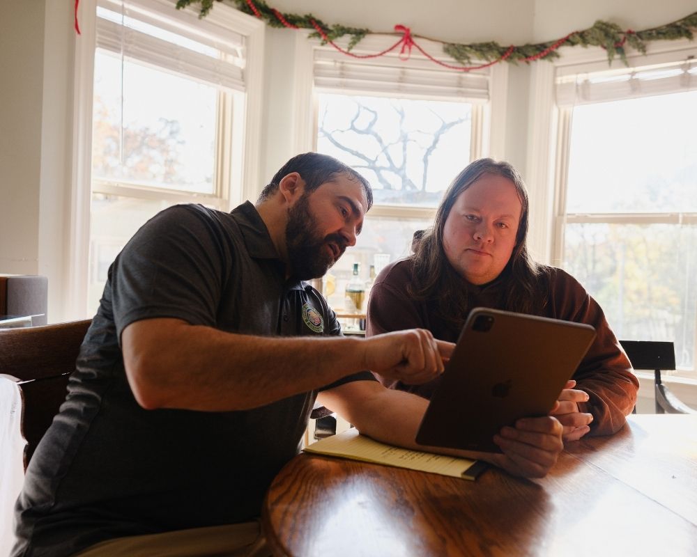 Two people sit at a wooden table by bright windows, looking at a tablet. One person points at the screen while explaining something to the other. Holiday garland decorates the windows in the background.