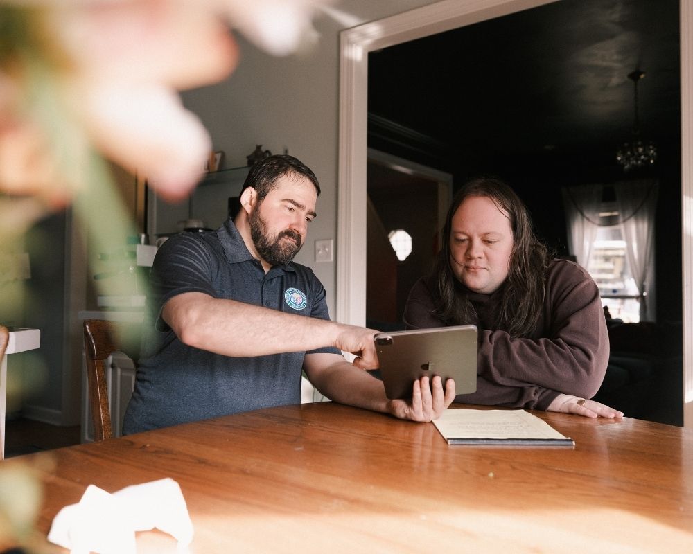 Two people sit at a wooden table indoors, looking at a tablet together. One person is pointing at the screen, and a notepad lies on the table beside them. The setting is warmly lit with a homey atmosphere.