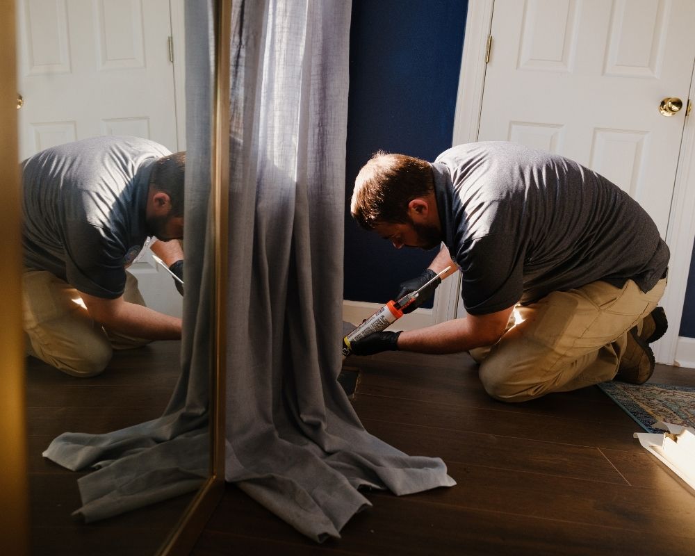 A man kneels on a wooden floor near a window, applying caulk along the baseboard with a caulking gun. Sunlight filters in, and a gray curtain partially covers the window.