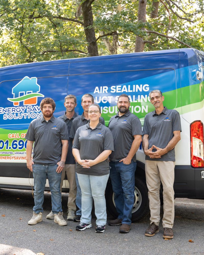 Six people wearing gray polo shirts stand in front of a branded Energy Saving Solutions van, smiling. The van is parked outdoors with trees in the background on a sunny day.