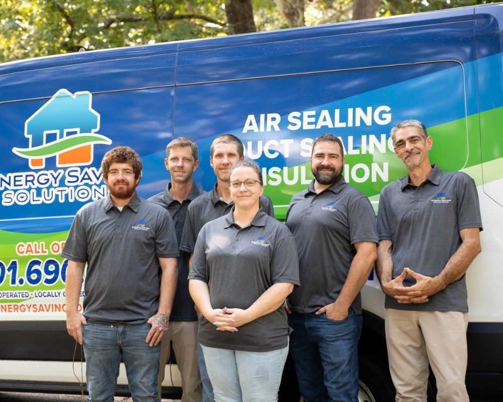 Six people in matching gray polos stand smiling in front of a blue and green van with the text Energy Saving Solutions and Air Sealing, Duct Sealing, Insulation visible on the side.